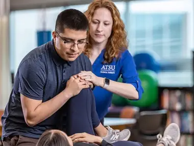 Doctor of Physical Therapy instructor demonstrating a leg stretch technique to a DPT student on a patient lying on a table.