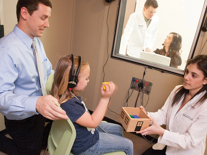 Young girl wearing headphones during a hearing test in an ATSU Audiology clinic, with a doctor conducting the test and her father observing