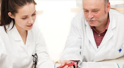 Young female osteopathic medical student in white coat under the tutelage of a seasoned clinician who is holding a red pen.
