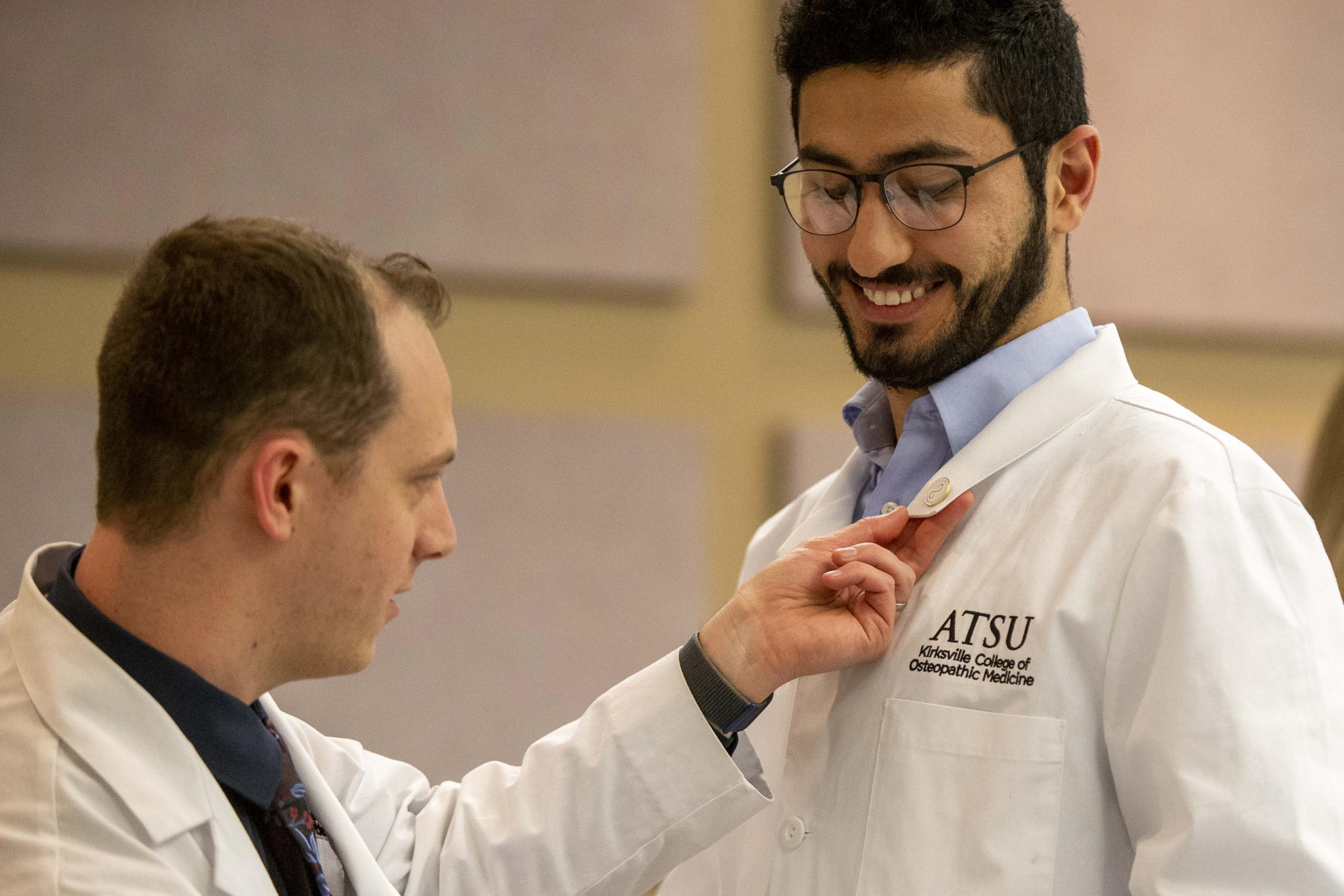 Two ATSU-KCOM students participate in a pinning ceremony. One student is looking down at the pin being placed on his lapel, while the other student holds the pin steady.