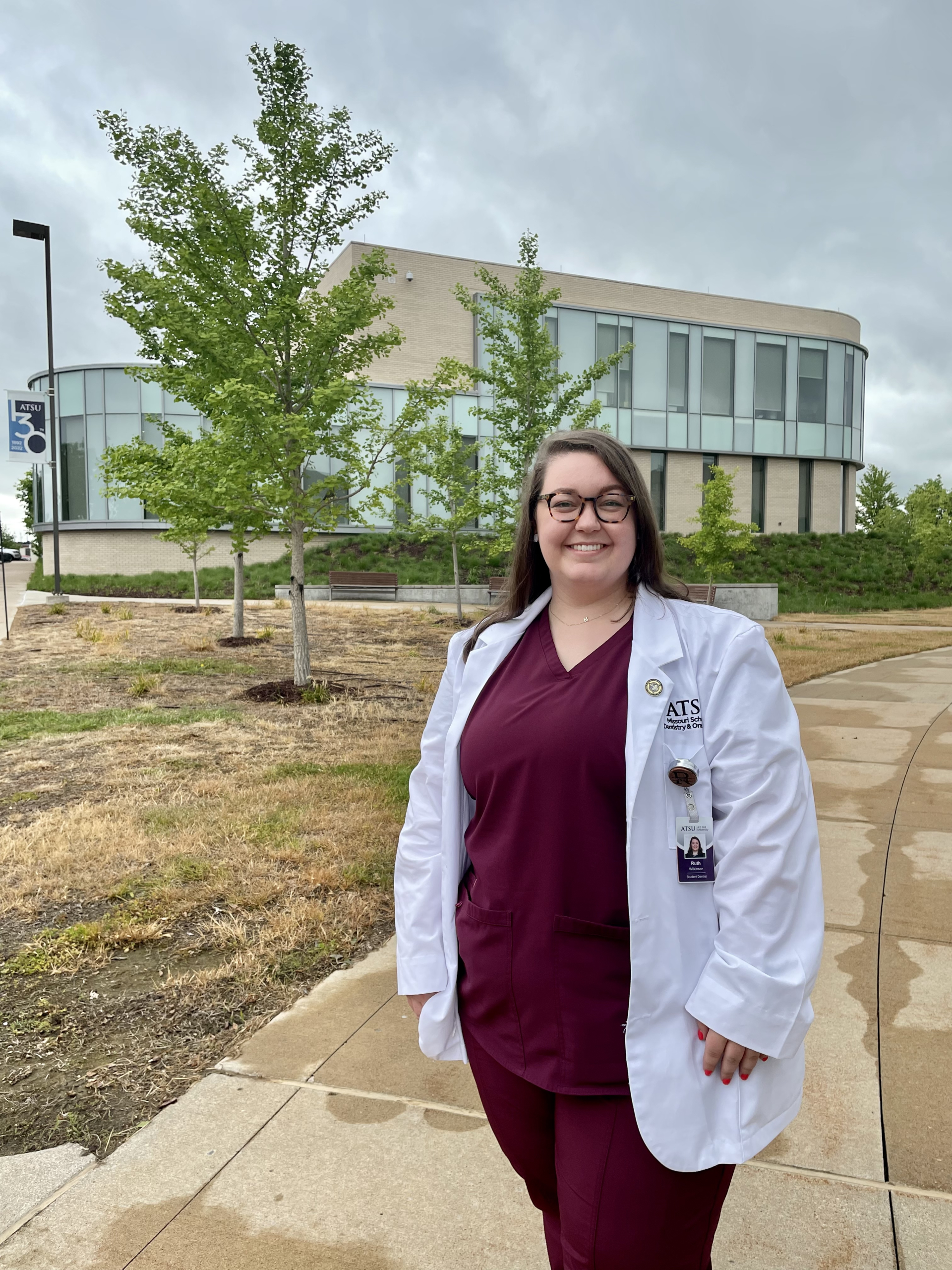 A dental student stands on a sidewalk outside of a building with large glass windows.