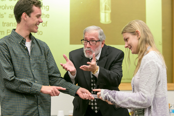Dr. Goldman stands between two students with his hands held up, as he performs a magic trick. The two students face each other holding an object between them.