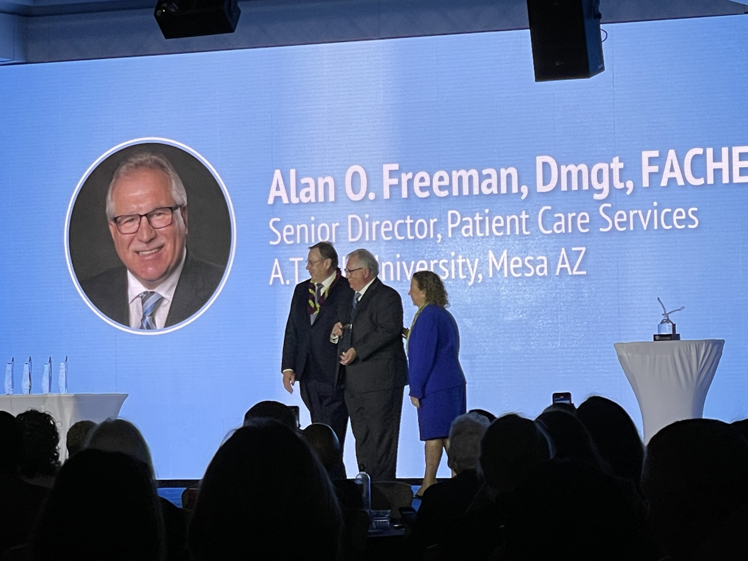 People are pictured on stage. The award recipient's photo and name appear on a large screen behind them.