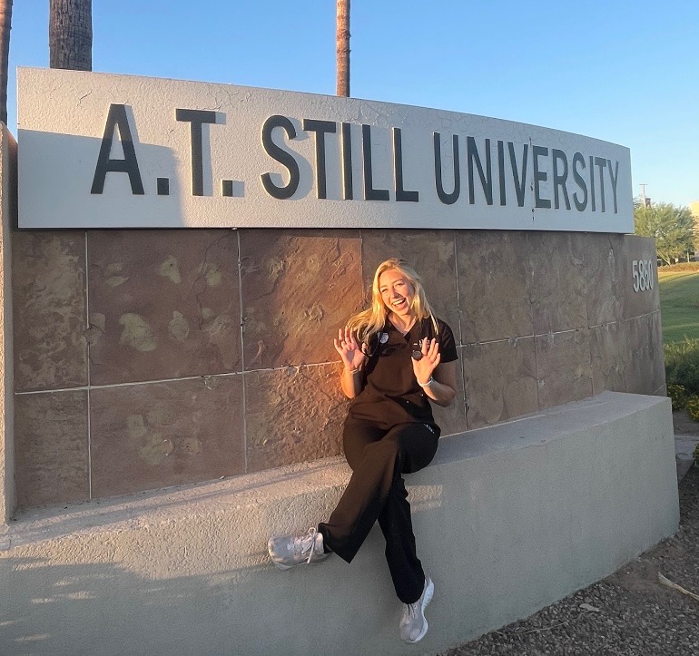 A student smiles as she sits in front of a large, stone A.T. Still University sign.