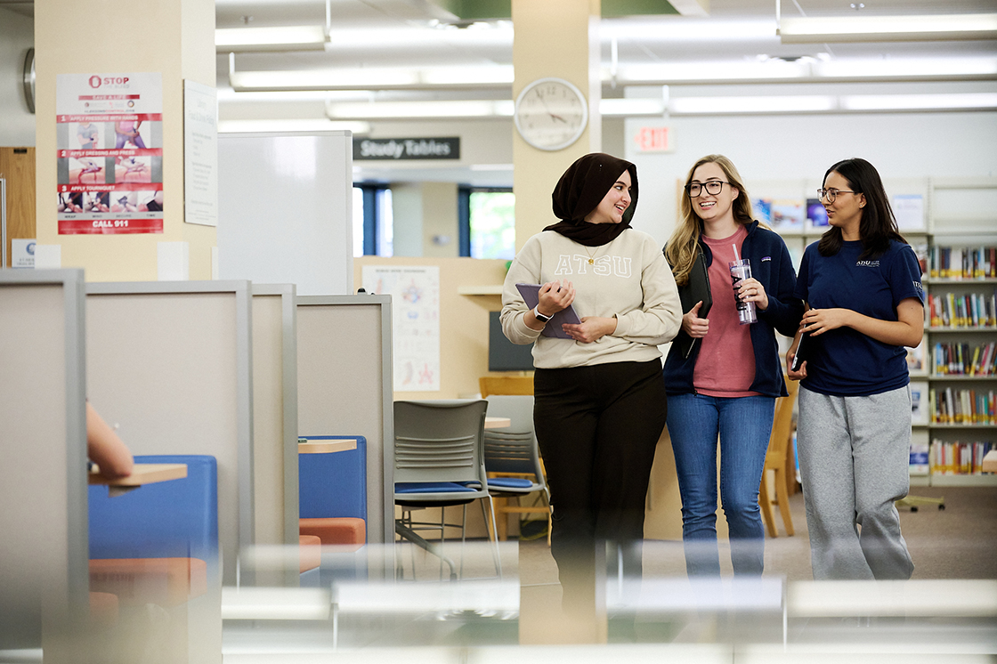 Three students walk through a library. Study tables are visible to the side, with library shelves in the background.