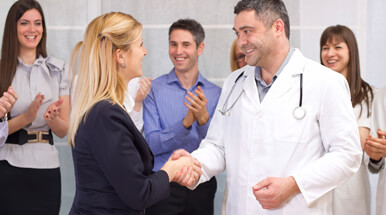 Male doctor in white lab coat shaking hands with a blonde female in a black suit.